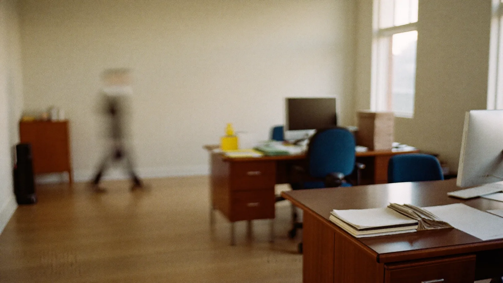 An office with wooden desks, a monitor and stacked paperwork, one person walking past the far wall, motion-blurred.