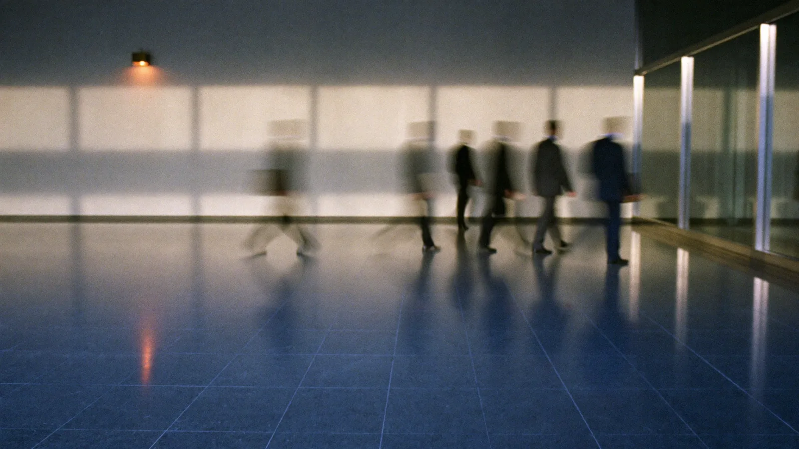 A line of people in suits carrying cases across a polished stone lobby past frosted glass, motion-blurred.