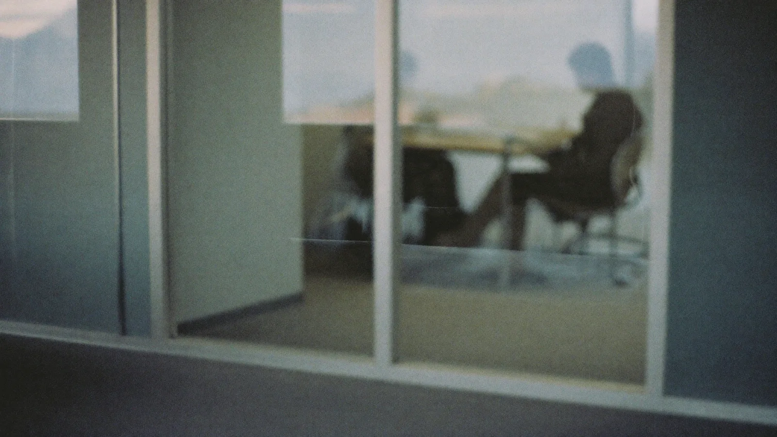 Two people at a boardroom table seen from outside through office glass partitions at dusk, heavily defocused.
