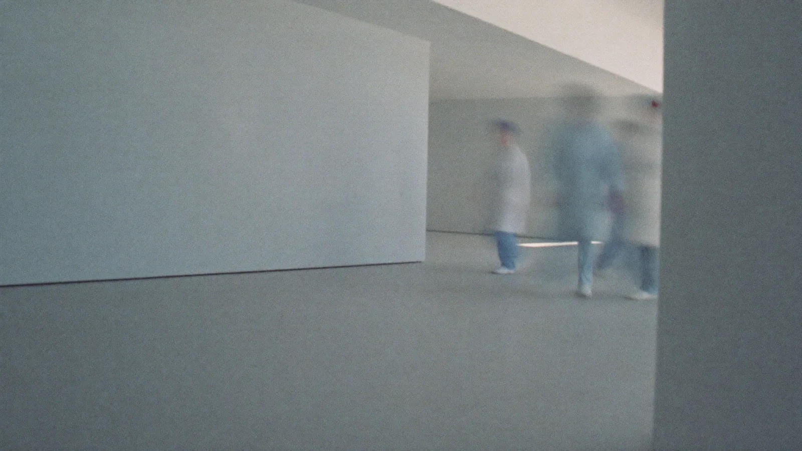 A bright hospital corridor, clinical staff in scrubs and a white coat pushing a trolley past a blank wall, motion-blurred.