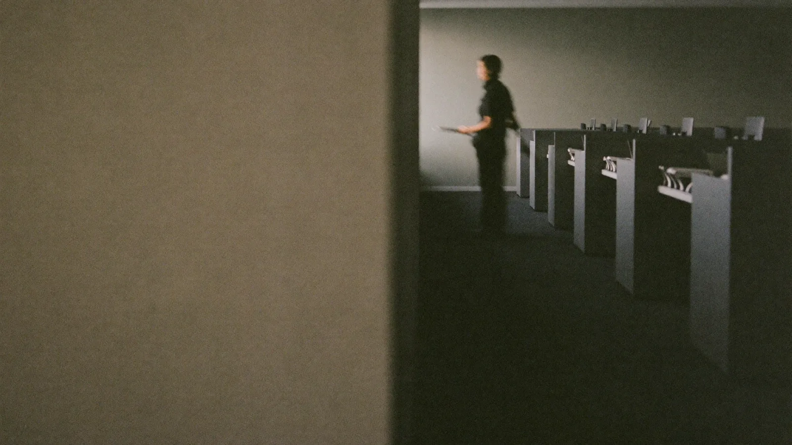 A dim open-plan office of empty desk partitions, one person standing at the end of the row holding papers.