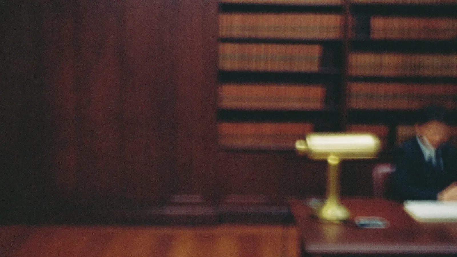 A wood-panelled library with shelves of bound volumes, a brass desk lamp, and a figure reading at a table, defocused.