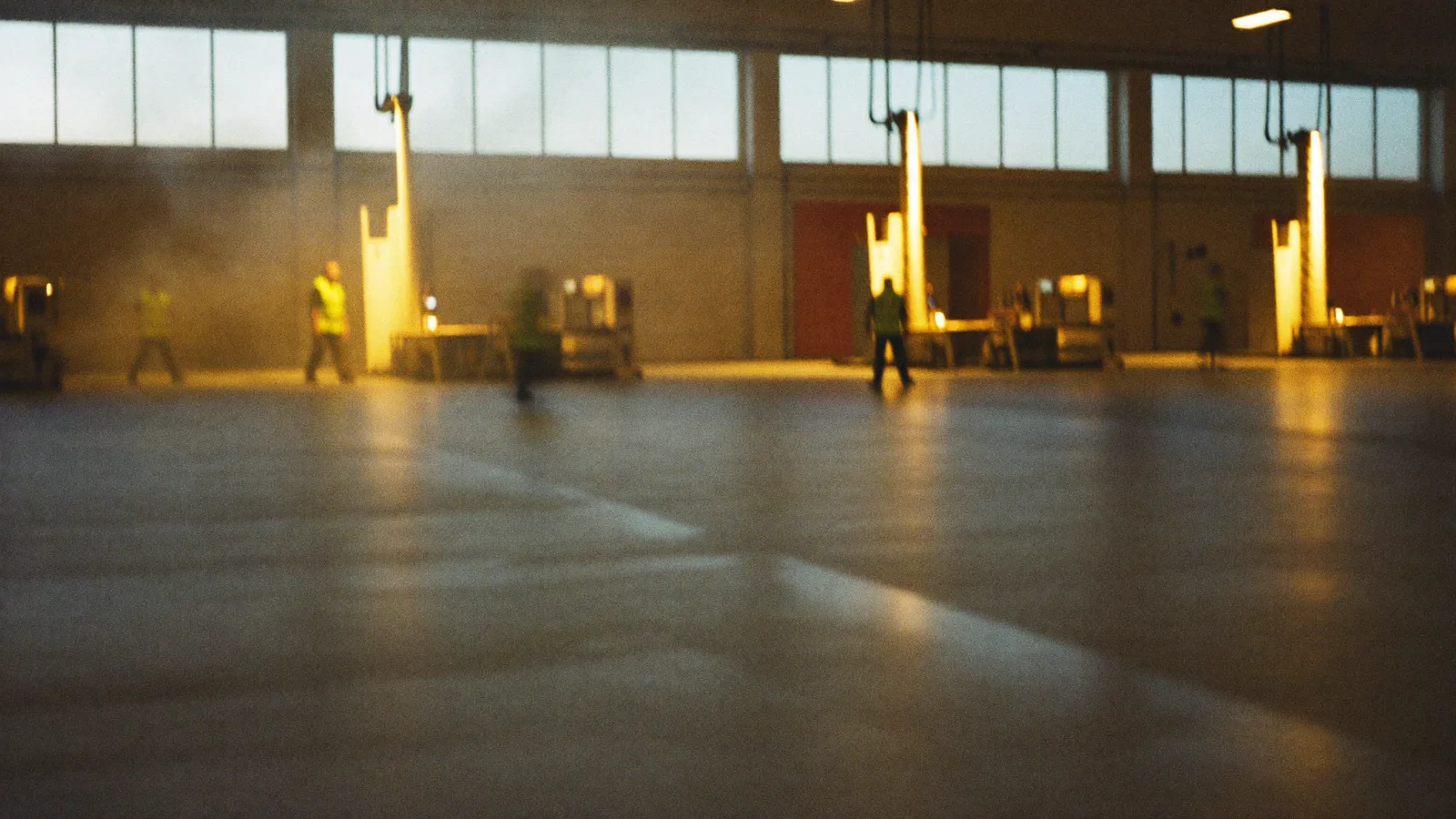 A large factory floor in low sun, workers in hi-vis walking between pallet stacks and machinery, motion-blurred.